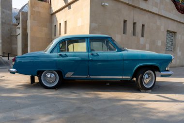 Bir binanın önünde mavi bir vintage araba park edilmiştir. Araba eski ve klasik bir tasarımı var. Sahne nostaljik ve eski bir atmosfer yayıyor, sanki geçmişte kalmış gibi.