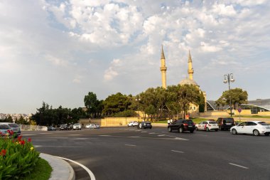 Arkasında büyük bir bina olan işlek bir cadde. Gökyüzü bulutlu ve güneş parlıyor