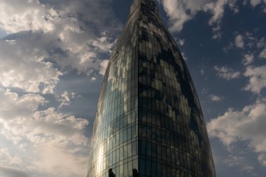 Bulutlu bir gökyüzüne karşı cam cepheli uzun bir bina görülür. Bina çok yüksek ve benzersiz bir şekli var. Gökyüzü parçalı bulutlu, mavi ve beyaz bulutlarla kaplı.