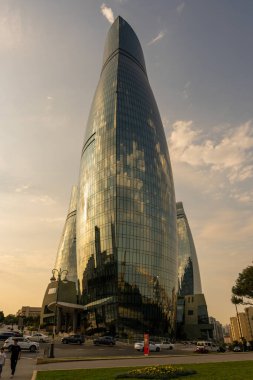 Bir sürü penceresi ve yan tarafında gökyüzünün yansıması olan yüksek bir bina. Gökyüzü bulutlu ve güneş batıyor.