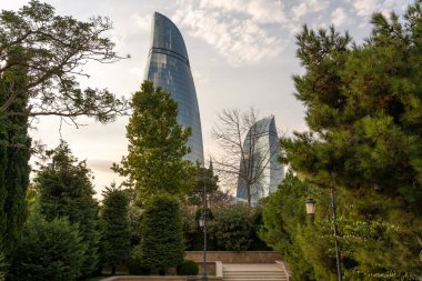 Arkasında büyük bir bina olan bir park. Bina uzun ve benzersiz bir şekli var. Gökyüzü bulutlu ve güneş batıyor. Parktaki ağaçlar yeşil ve huzurlu bir atmosfer sağlıyor.