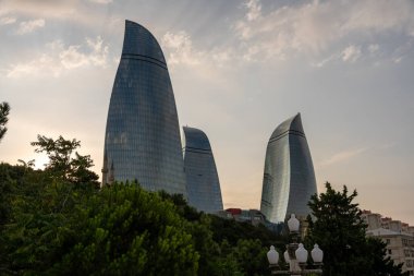 Bir sürü penceresi ve arka planda yeşil parkı olan üç büyük bina. Gökyüzü bulutlu ve güneş batıyor.
