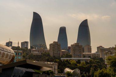 Arka planda şehir silueti olan üç büyük bina. Binalar camdan yapılmış ve eşsiz bir dizaynı var. Gökyüzü bulutlu, ortama biraz karamsar bir hava katıyor.