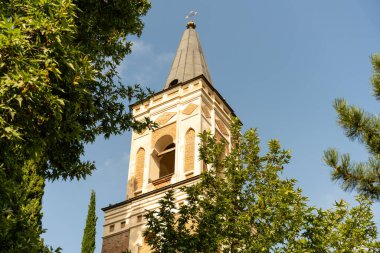 Sivri çatılı ve tepesinde haç olan uzun tuğla bir kule. Kule ağaçlarla çevrili ve arka planda açık mavi bir gökyüzü var.