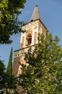 Sivri uçlu ve küçük pencereli uzun tuğlalı bir kule. Kule ağaçlarla çevrili ve gökyüzü temiz.
