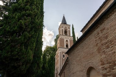 Önünde uzun bir kule ve büyük bir ağaç olan tuğla bir bina. Ağaç yeşil ve bina kahverengi.