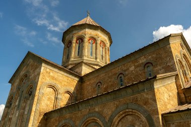 Çan kulesi ve tepesinde haç olan büyük bir bina. Bina kahverengi ve bir sürü penceresi var. Gökyüzü mavi ve bazı bulutlar var.