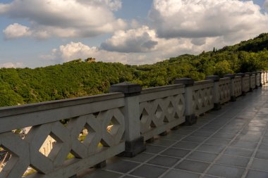 Korkuluklu ve orman manzaralı taş bir duvar. Tırabzan taştan yapılmış ve elmas deseni var. Ormanın manzarası güzel ve huzurlu.