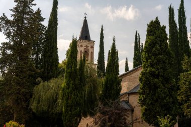 Uzun bir çan kulesi ve arka planda büyük bir ağaç grubu olan bir kilise. Ağaçlar yeşil ve doludur. Huzurlu ve huzurlu bir atmosfer yaratırlar.