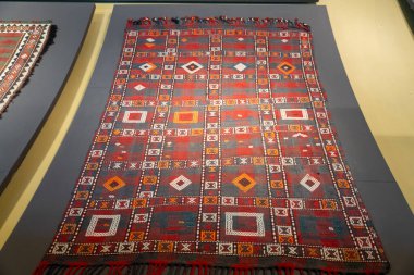 Duvarda kare ve üçgen desenli bir halı sergileniyor. Halı mavi ve kırmızı, beyaz kenarlı.