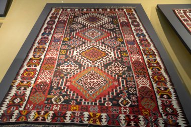 Duvarda kırmızı ve beyaz desenli bir halı asılı. Halı büyük ve rengarenk, kırmızı, beyaz ve sarı karışımı. Tasarım karmaşık ve detaylı, birçok küçük kare ve üçgenle