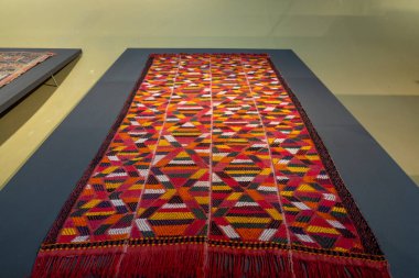 Bir masanın üzerinde geometrik desenli renkli bir halı sergilenir. Halı bir müzede ya da galeride sergileniyor.