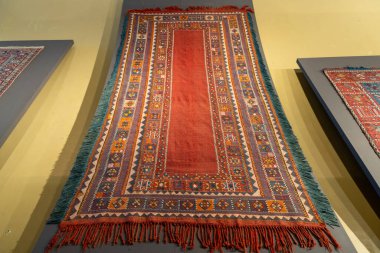 Duvarda mavi çizgili bir halı sergilenir. Halı kırmızı ve mavi, yeşil çizgili.