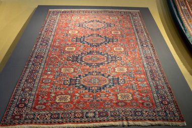 Mavi kenarlı kırmızı ve mavi bir halı. Halı sergileniyor.