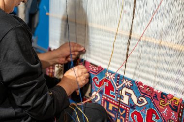 Bir adam yerde oturuyor ve bir halı üzerinde çalışıyor. Halı mavi ve beyaz ve kırmızı ve sarı bir deseni var. Adam halıyı dikmek için iğne iplik kullanıyor. Huzur dolu bir sahne.