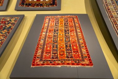 Sarı kenarlı bir müzede bir halı sergileniyor. Halı renkli ve üçgen bir deseni var.