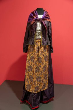 Bir kadın mor ve turuncu bir elbise ve mor bir eşarp giyiyor. Kadın kırmızı bir arka planın önünde duruyor.