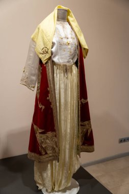 Bir kadın sarı atkılı kırmızı beyaz bir elbise giyiyor. Elbise uzun ve zarif, ve eşarp kıyafete renk katıyor. Sahne entelektüellik ve zarafettir.