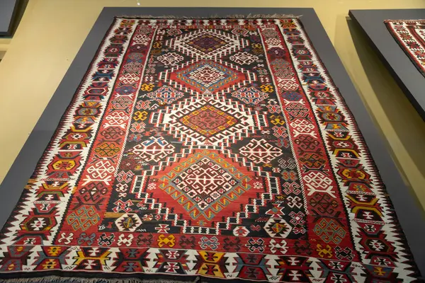 Duvarda kırmızı ve beyaz desenli bir halı asılı. Halı büyük ve rengarenk, kırmızı, beyaz ve sarı karışımı. Tasarım karmaşık ve detaylı, birçok küçük kare ve üçgenle