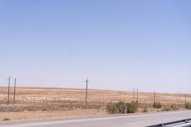Birkaç ağaç ve elektrik hattı olan çorak bir çöl manzarası. Gökyüzü açık ve mavi. Manzara ıssız ve yalnız.