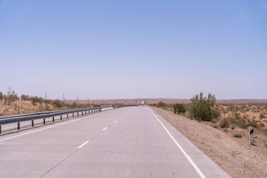Üzerinde mavi bir gökyüzü olan uzun, boş bir yol. Yol uzun, kuru otlarla kaplı ve üzerinde ne araba ne de insan var.