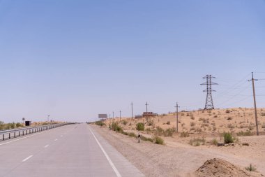 Arka planda elektrik hattı olan uzun bir yol. Gökyüzü açık ve güneş parlıyor.