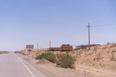 İlan panosu ve uzakta küçük bir bina olan bir yol. Gökyüzü açık ve güneş parlıyor.