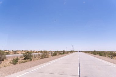 Üzerinde açık mavi gökyüzü olan uzun, boş bir yol. Yol ağaçlarla kaplı ve yolun ortasında bir direk var.