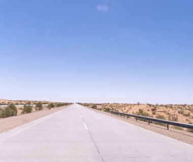 Üzerinde açık mavi gökyüzü olan uzun, boş bir yol. Yol betondan yapılmıştır ve bir çöl manzarasıyla çevrilidir.