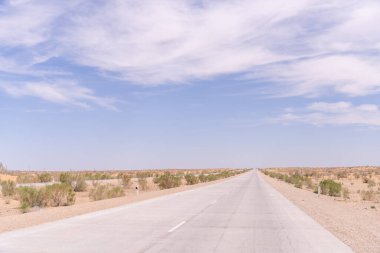 Üzerinde açık mavi gökyüzü olan uzun, boş bir yol. Yol bir çöl manzarasıyla çevrilidir.