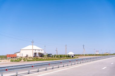 Kırsal bir yol, beyaz bir ev ve birkaç bina daha. Gökyüzü açık ve mavi