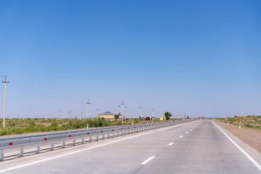 Üzerinde mavi bir gökyüzü olan uzun, boş bir yol. Yolda hiç araba yok ki. Yol uzun metal direklerle kaplı.