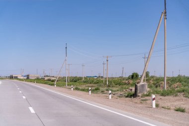 Arka planda birkaç ağaç ve kablolu birkaç direk olan bir yol. Gökyüzü mavi ve berrak