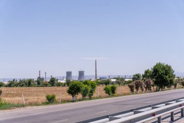 Yan tarafında ağaç ve arka planında elektrik santrali olan bir yol. Gökyüzü açık ve güneş parlıyor.