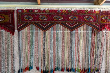 Duvarında boncuklu renkli bir halı asılı duruyor. Halı farklı renklerde ipliklerden yapılmış ve eşsiz bir tasarımı var.