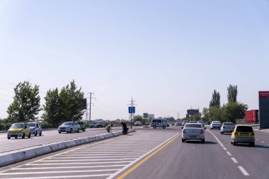 Arabaları olan kalabalık bir cadde ve kaldırımda yürüyen yeşil gömlekli bir adam. Sahne meşgul ve hareketli.