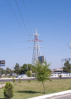 Uzun bir elektrik hattı kulesi yolun yanındaki çimen tarlasında duruyor. Gökyüzü açık ve mavi, ve yol boyunca park edilmiş birkaç araba var.