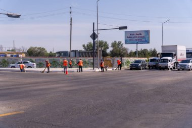 Bir grup inşaat işçisi bir caddede duruyor. İşçiler turuncu yelek giyiyorlar ve bir yol üzerinde çalışıyorlar. Yolun kenarında park edilmiş birkaç araba var.