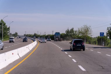 Yoğun bir otoyol ve yolda giden bir araba. Yolda bir kamyon da dahil olmak üzere bir sürü araba var. Gökyüzü açık ve güneş parlıyor.