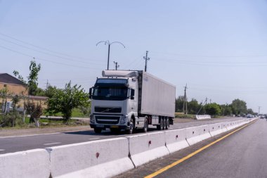Büyük beyaz bir kamyonet yolda ilerliyor. Kamyon bir otoyolda ve ağaçlarla çevrili. Gökyüzü açık ve güneş parlıyor.