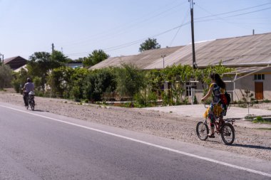Bir kadın, arkasında bisikletli bir adamla birlikte bisiklet sürüyor. Kadın sarı bir sırt çantası takıyor.