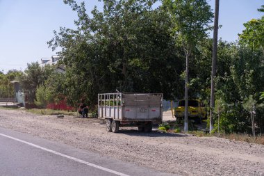 Yolun kenarına park edilmiş bir kamyon var. Bir adam kamyonun yanında yerde oturuyor.