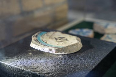 Kırık bir çömlek parçası mavi ve beyaz bir dizaynla siyah bir yüzeyde duruyor. Parça çatlamış ve kırılmış, ona bir tarih hissi veriyor ve aşınmış.