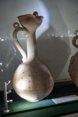 Kolu olan bir vazo sergileniyor. Vazo beyaz ve üzerinde bir desen var.