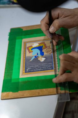 Bir insan yeşil bir kağıt parçasına resim çiziyor. Çizimde mavi gömlekli ve mavi şapkalı bir kişi var. Elinde bir cetvel ve siyah bir kalem var.