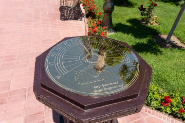 Parktaki ahşap bir üste büyük, süslü bir saat duruyor. Saat kırmızı çiçekler ve otlarla çevrili. Huzurlu ve huzurlu bir atmosfer yaratıyor.