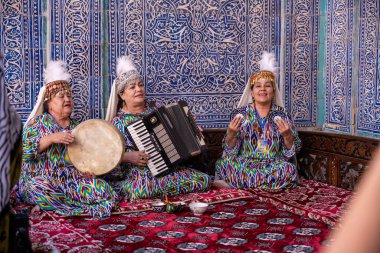 Mavi bir duvarın önünde enstrüman çalan üç kadın. Kadınlar renkli kıyafetler giyip klavye ve davul çalıyorlar.
