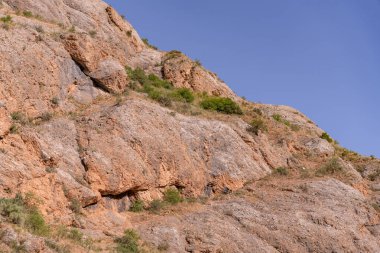 Arka planda mavi gökyüzü olan kayalık bir yamaç. Kayalar kahverengi, çimenler yeşil.