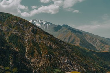 Dağlar yeşil ağaçlar ve kahverengi kayalarla kaplıdır. Gökyüzü bulutlu ve güneş parlıyor