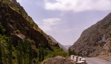 İçinden yol geçen bir dağ sırası. Yol ağaçlar ve kayalarla kaplı. Gökyüzü açık ve güneş parlıyor.